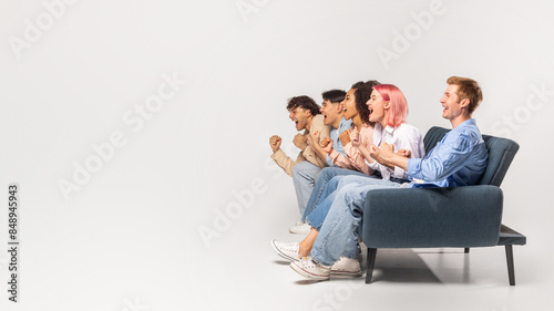 Five diverse young friends are sitting on a couch in a studio setting. They are all looking off to the side, cheering excitedly. They are likely watching a sporting event or a game show, copy space