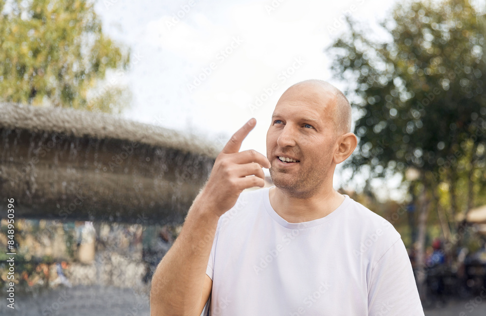Bald man in white t-shirt pointing upwards and smiling near outdoor fountain Bald man in white t-shirt pointing upwards and smiling near outdoor fountain