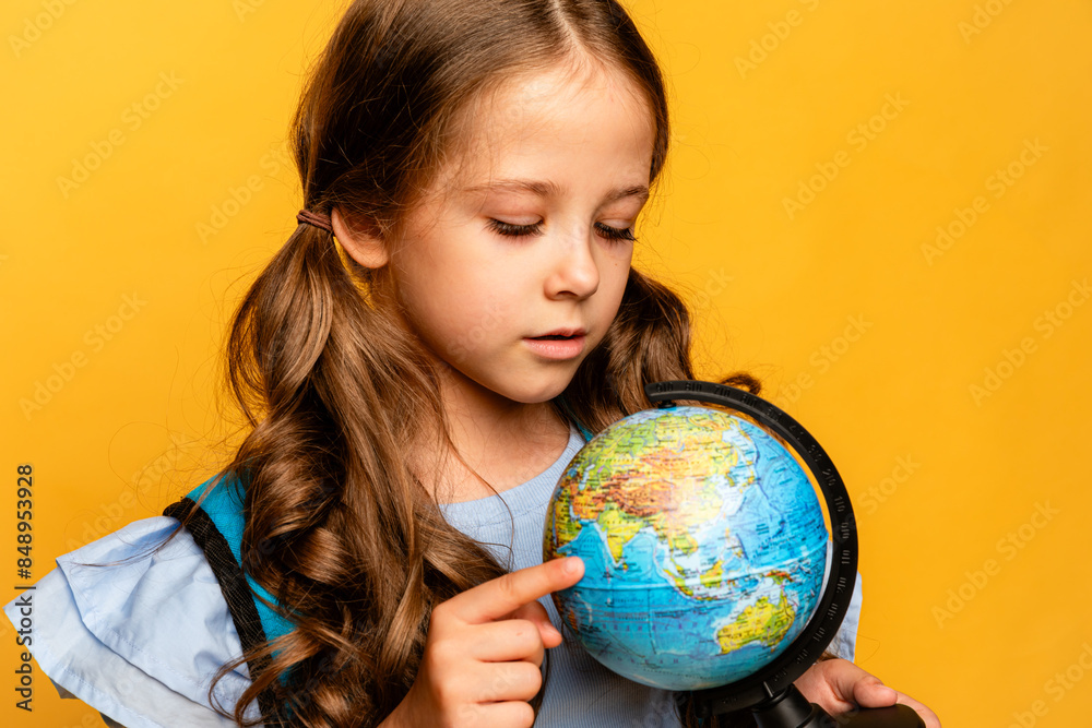 Happy first year schoolgirl girl in a blue t-shirt and jeans on a yellow background with a globe in her hands and backpack. Copy space. Back to school concept. Happy first year schoolgirl girl in a blue t-shirt and jeans on a yellow background with a globe in her hands and backpack. Copy space. Back to school concept.