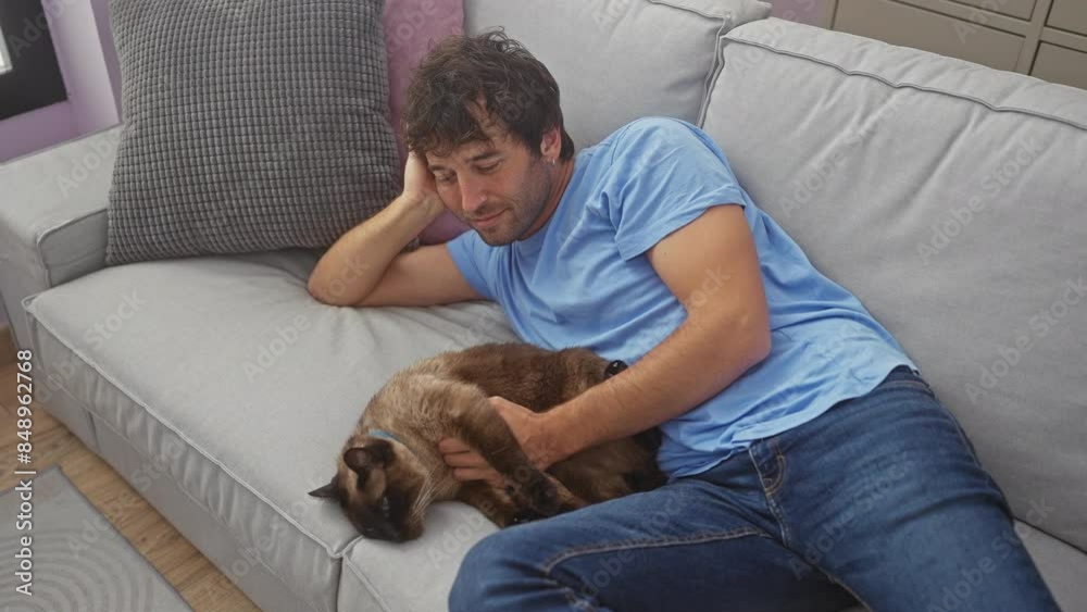 A relaxed young man in a blue shirt cuddles with his siamese cat on a grey sofa indoors. A relaxed young man in a blue shirt cuddles with his siamese cat on a grey sofa indoors.