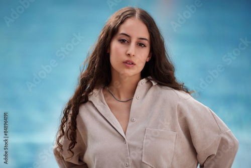 Portrait of a young woman with brown long hair and brown shirt, blouse, looking into the camera. Blue water in the background. 