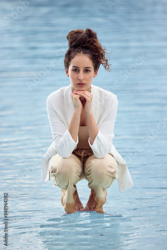Full body fashion portrait of a young beautiful woman in the water with brown hair, beautiful skin, white blazer and brown trousers. Model in the pool looking into the camera. On blue Background.