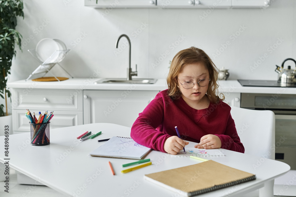 A little girl with Down syndrome colors at a table in her home kitchen. A little girl with Down syndrome colors at a table in her home kitchen.