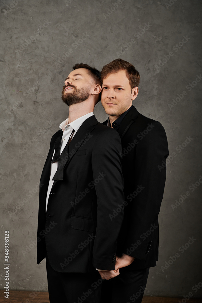 Two men in elegant suits stand close together against a gray backdrop. Two men in elegant suits stand close together against a gray backdrop.