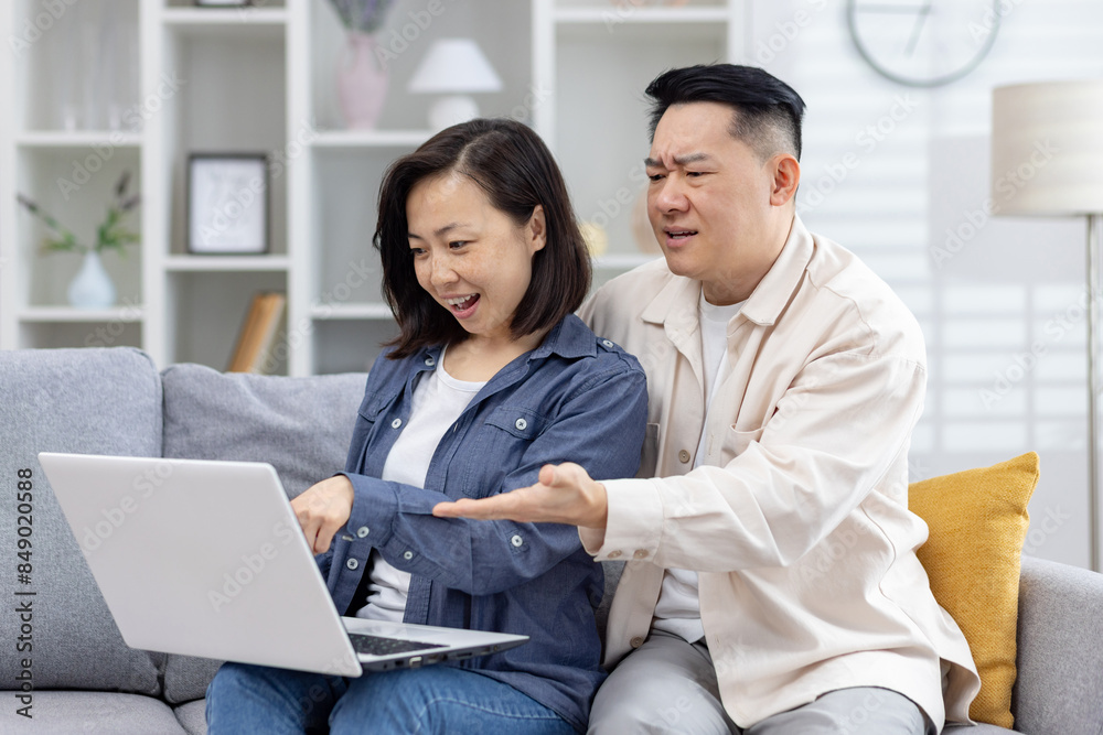 Asian couple having a disagreement over online shopping while sitting on a sofa at home. The woman is excited while the man seems concerned. Asian couple having a disagreement over online shopping while sitting on a sofa at home. The woman is excited while the man seems concerned.