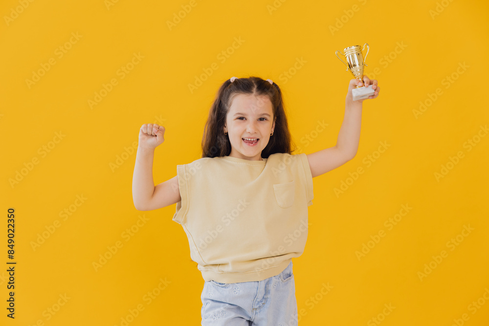 Happy little girl with trophy shows a golden cup and success isolated on a yellow background Happy little girl with trophy shows a golden cup and success isolated on a yellow background