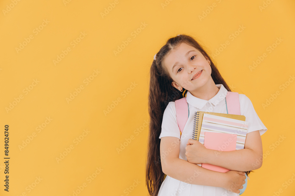 Cute little first grader girl in white school uniform with backpack holding notebooks and books on yellow background Cute little first grader girl in white school uniform with backpack holding notebooks and books on yellow background