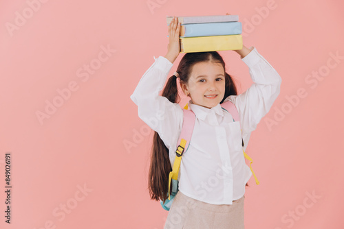 Cute smiling child school girl in uniform with backpack holding books on her head on pink background