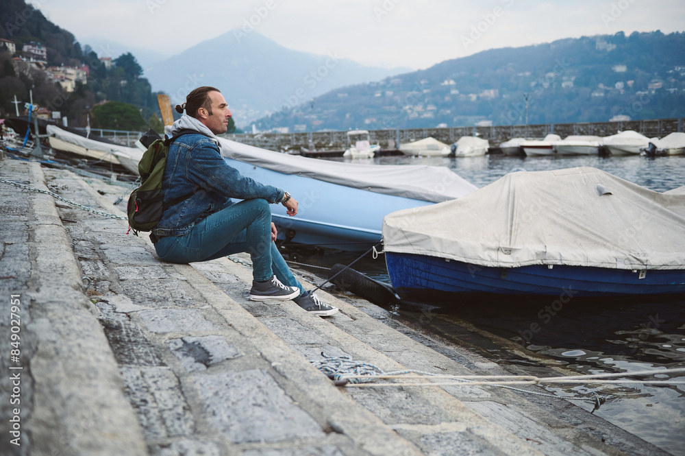 Fototapeta premium Man sitting on a dock by a marina with mountains in the background