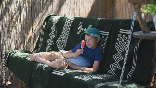 Cute pre-teen boy in green panama eating ice cream while swinging on a swing outdoors photo