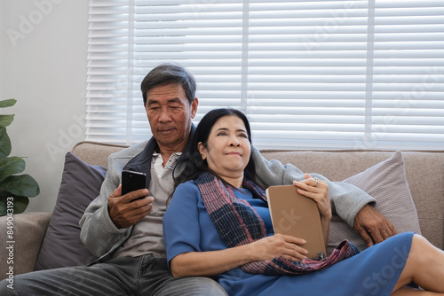 Elderly Couple Relaxing in Modern Living Room, Enjoying Leisure Time Together on Comfortable Sofa, Using Digital Devices, and Embracing a Peaceful and Cozy Home Environment