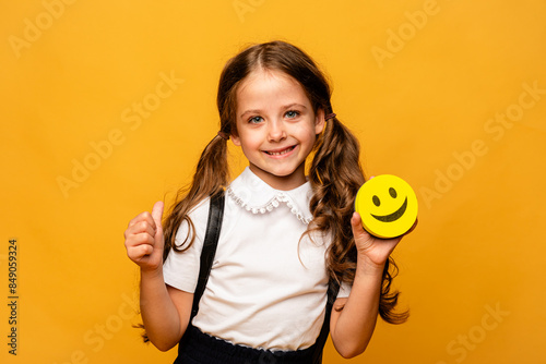 A funny schoolgirl is holding a happy emoji emotion icon in her hands and showing thumbs up on yellow background. International World smile Day. World emoji day. School Birthday Party. Emotions. photo