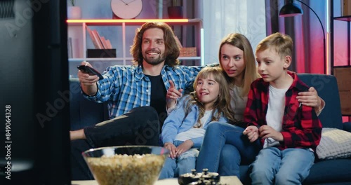 Happy family man woman and kids watching comedy or entertaining show gathering together in front of TV set at home in the evening. Leisure concept.