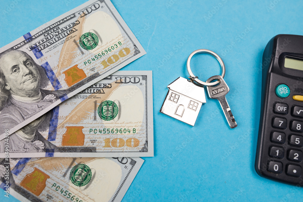 A small house with keys on a background of hundred dollar bills and a calculator. A small house with keys on a background of hundred dollar bills and a calculator.