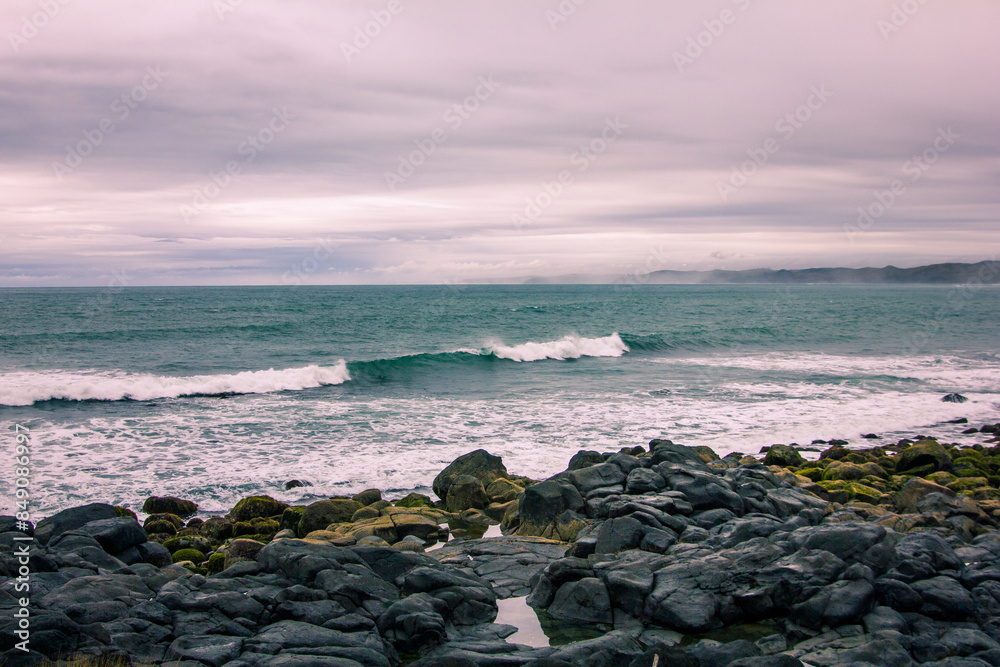 Large waves crash into with coastal rocks covered in green moss. Dramatic sea coast background. Raglan, New Zealand Large waves crash into with coastal rocks covered in green moss. Dramatic sea coast background. Raglan, New Zealand