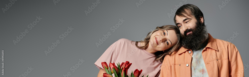 A man and a woman in sophisticated attire pose together, with the woman holding a bouquet of red tulips. A man and a woman in sophisticated attire pose together, with the woman holding a bouquet of red tulips.
