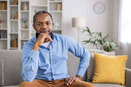 Confident man sitting on a couch in a cozy, modern home interior. He is smiling and looking relaxed, conveying a sense of comfort and ease.