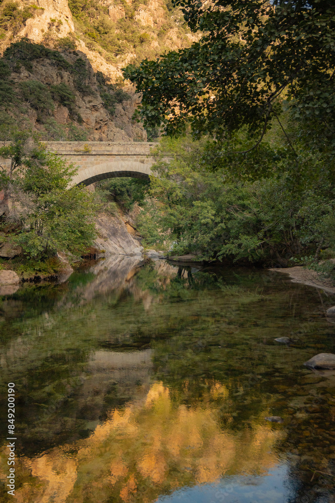 Fototapeta premium Reflection of mountain on the surface of a river flowing through a canyon with ancient stone bridge crossing