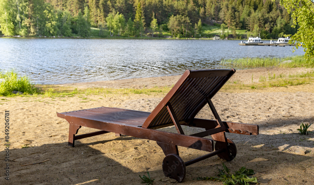A wooden lounge chair sits on a sandy beach, with a view of a calm lake and a forested shoreline. The sun casts long shadows on the sand, creating a serene atmosphere. A wooden lounge chair sits on a sandy beach, with a view of a calm lake and a forested shoreline. The sun casts long shadows on the sand, creating a serene atmosphere.