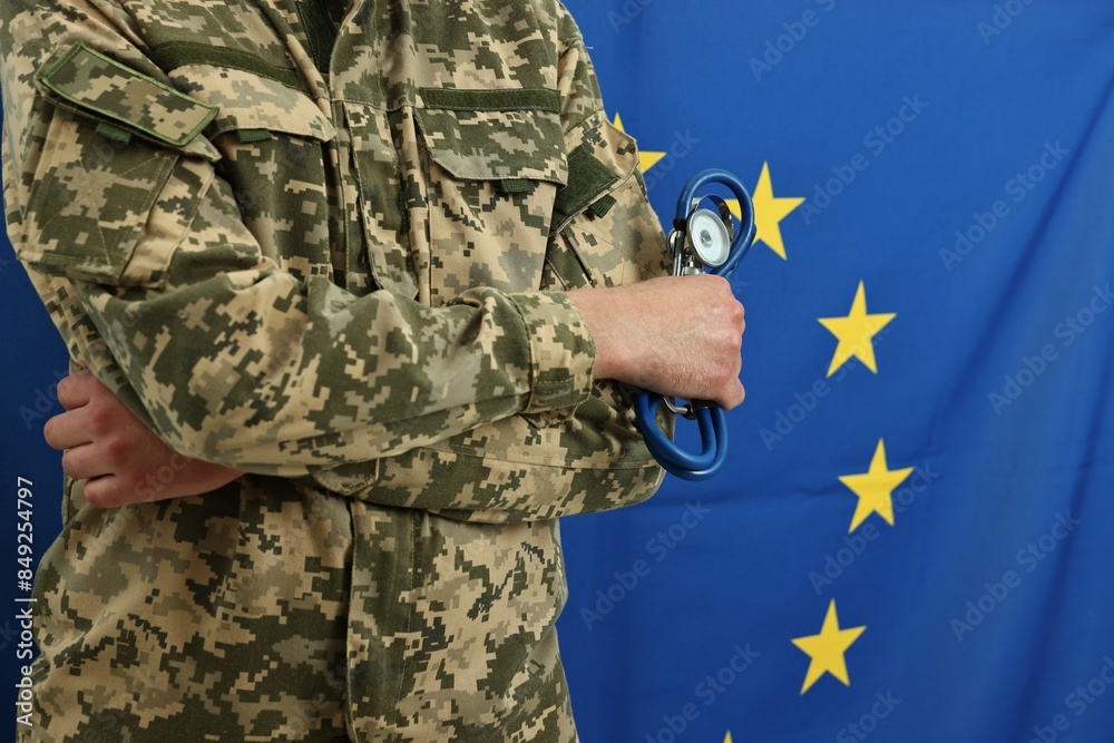 Fototapeta premium Man in military uniform with crossed arms and stethoscope against flag of European Union, closeup. Health care concept