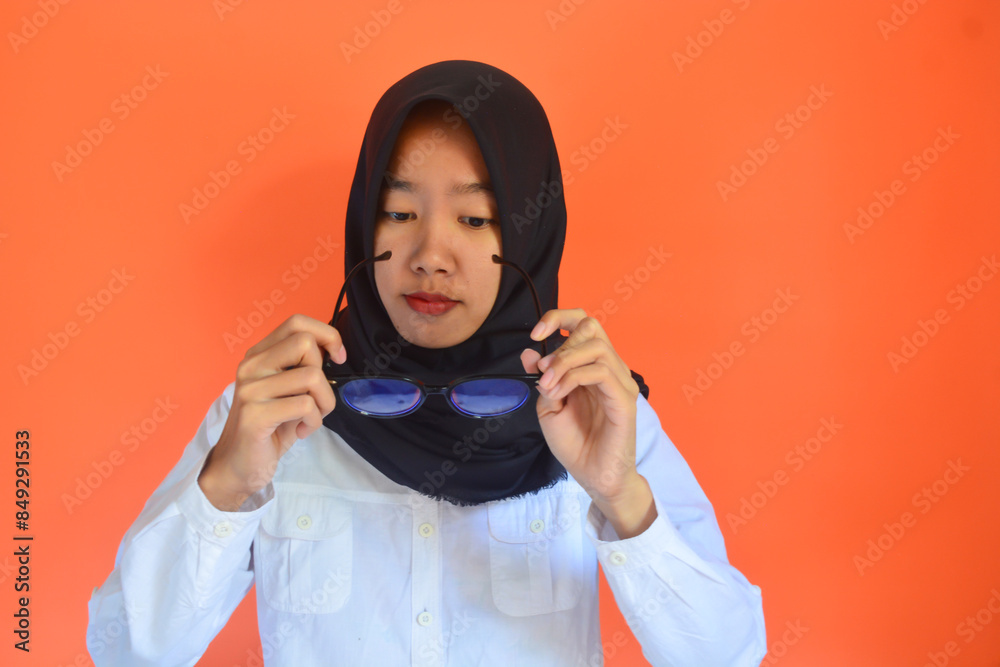 Beautiful young Asian business woman wearing white shirt and black hijab smiling, showing, with raised hand gesture and wearing glasses isolated on orange background Beautiful young Asian business woman wearing white shirt and black hijab smiling, showing, with raised hand gesture and wearing glasses isolated on orange background