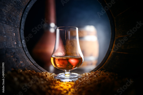 Tasting glass with whiskey standing inside of old oak barrel filled with barley grains. Traditional alcohol distillery concept photo