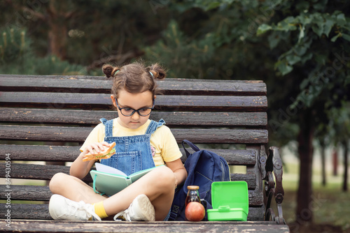Cute little schoolgirl eating from lunch box outdoor sitting on a bench. Food for kids.