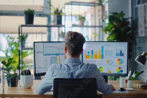 Young professional analyzing financial data on multiple computer screens in a modern office with plants in the background.