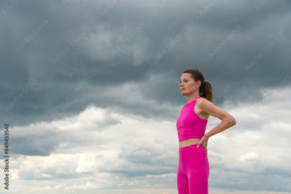 Sporty woman standing resting with hands on hips wearing pink sports clothing against a dramatic sky. Sporty woman standing resting with hands on hips wearing pink sports clothing against a dramatic sky.