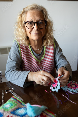 blonde woman in her sixties in a checkered crochet dressing gown, red and gel nails. portrait looking at the camera and smiling.