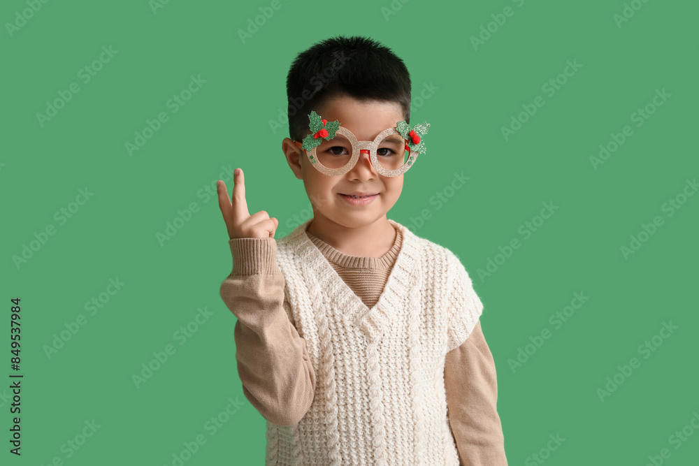 Cute little boy in paper eyeglasses and vest showing victory gesture on green background Cute little boy in paper eyeglasses and vest showing victory gesture on green background