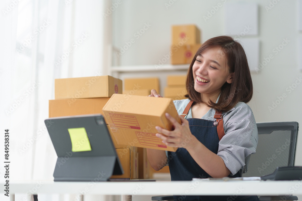 Smiling young woman in apron managing online order packaging in home office with computer on table and boxes around. Smiling young woman in apron managing online order packaging in home office with computer on table and boxes around.