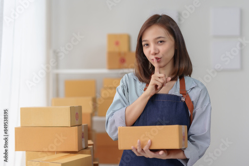 Schilderij op canvas Woman in apron holding a cardboard box with a finger to her lips in a quiet gesture in a room filled with stacked boxes