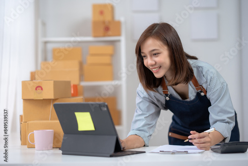 Young woman managing online business from home. She is working on a laptop surrounded by parcels, representing e-commerce and small business. © amnaj