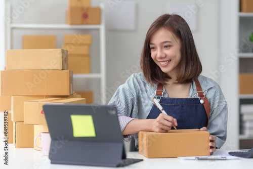 Young woman wearing an apron, packing a box while working from home with laptop and stack of cardboard boxes around her. © amnaj
