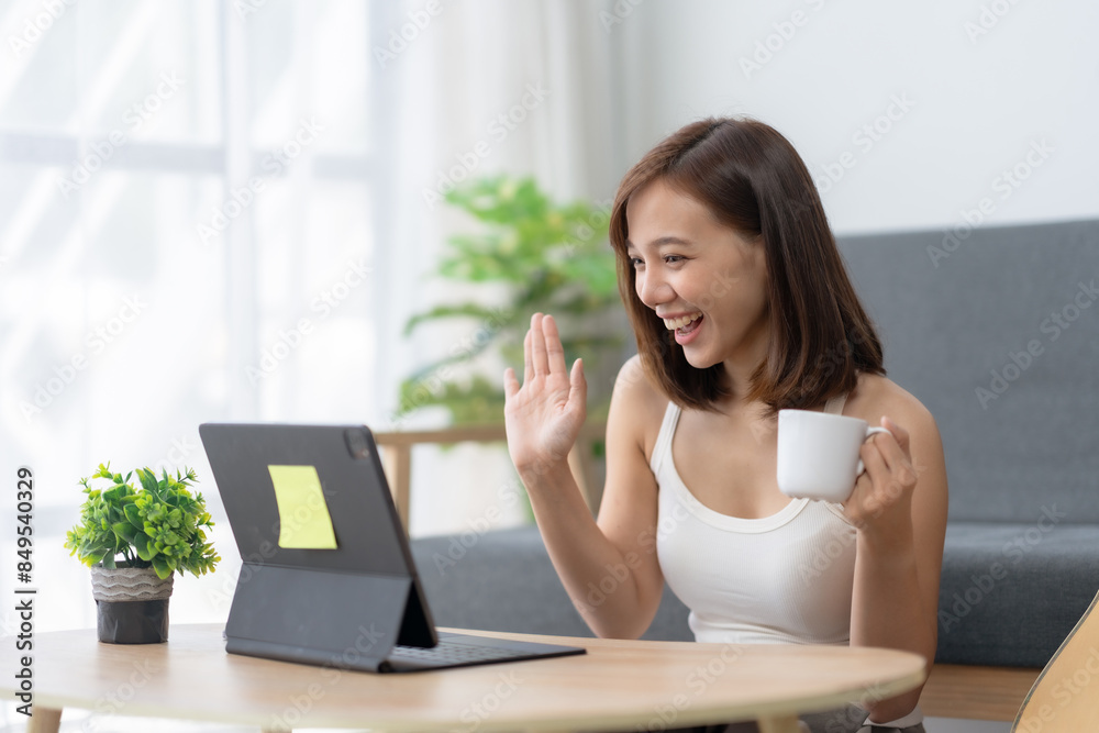 Happy woman having a video call, holding a cup of coffee and smiling, sitting at a table with a tablet at home. Happy woman having a video call, holding a cup of coffee and smiling, sitting at a table with a tablet at home.