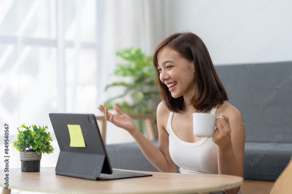 Young woman enjoying a video call on her tablet while holding a coffee cup, sitting comfortably indoors with a potted plant nearby. Young woman enjoying a video call on her tablet while holding a coffee cup, sitting comfortably indoors with a potted plant nearby.