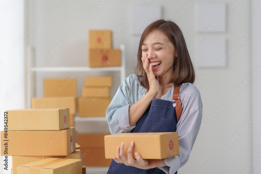 Smiling woman holding boxes in a home office, preparing for shipping with excitement and enthusiasm, surrounded by packed items.