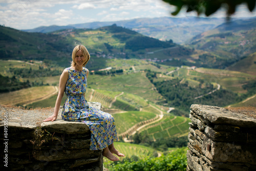 A woman in a light dress sits on a stone wall of the vineyards in the Douro Valley.