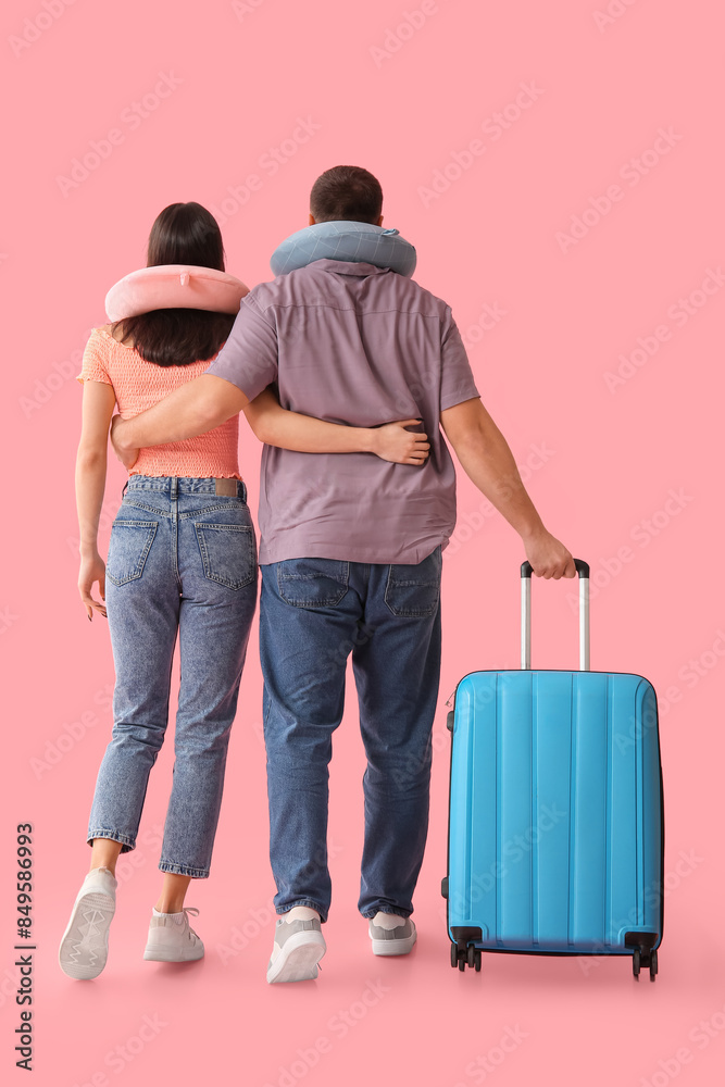 Beautiful young couple of tourists with suitcase and travel pillows on pink background, back view Beautiful young couple of tourists with suitcase and travel pillows on pink background, back view