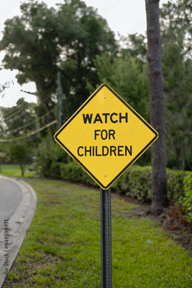 Close-up, crooked, WATCH FOR CHILDREN yellow, diamond sign with space on the left side and tree-lined road and fence in the background, verticle. Close-up, crooked, WATCH FOR CHILDREN yellow, diamond sign with space on the left side and tree-lined road and fence in the background, verticle.
