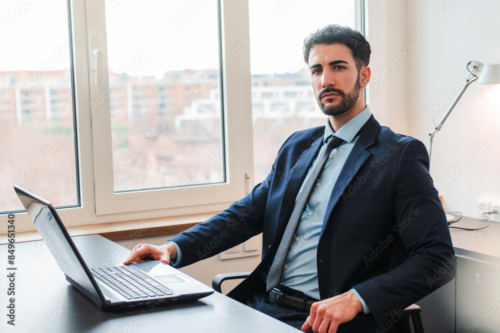 Individual portrait of one well dressed business man sitting at desk with a laptop and looking at camera with serious expression. Office worker staring front at workplace. Successful executive or ceo Individual portrait of one well dressed business man sitting at desk with a laptop and looking at camera with serious expression. Office worker staring front at workplace. Successful executive or ceo