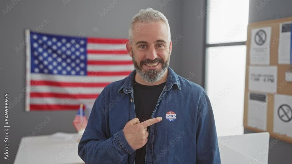 Caucasian man with beard indoors pointing at 'i voted today' sticker with american flag backdrop