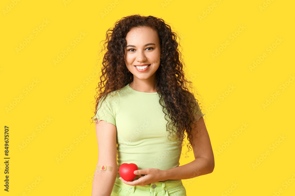 Young female African-American blood donor with applied medical patch and heart on yellow background Young female African-American blood donor with applied medical patch and heart on yellow background