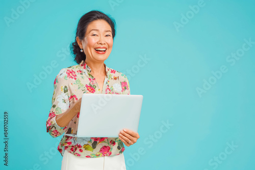 A middle-aged Asian woman in her forties, dressed casually in a floral blouse and white pants, standing against a light blue background, holding a laptop and looking excited. photo