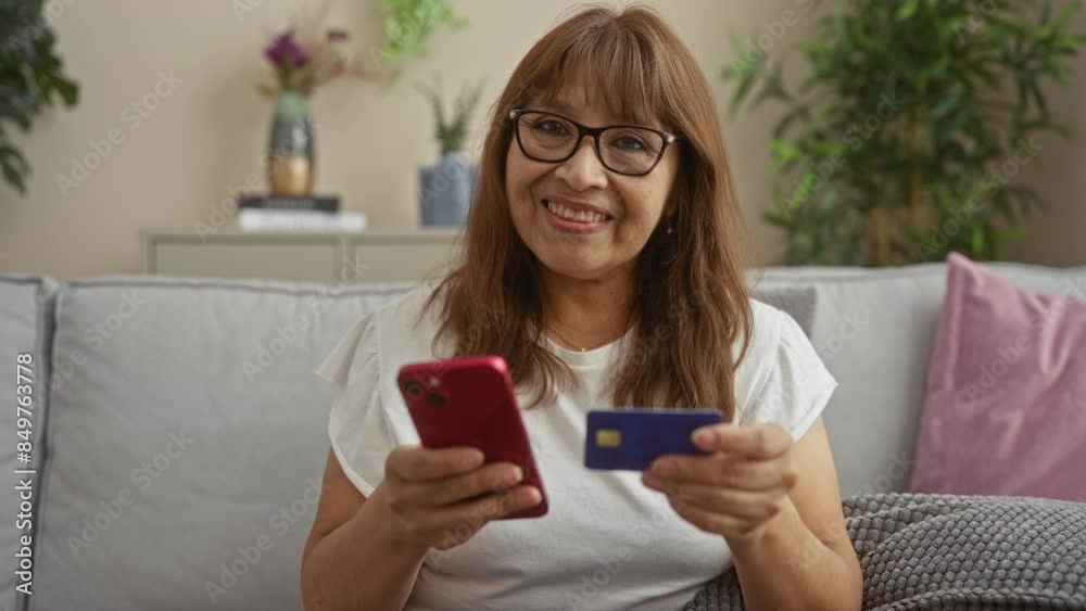 A mature woman sitting in her living room holding a smartphone and credit card, smiling while engaging in online shopping or banking, surrounded by cozy home decor and indoor plants. A mature woman sitting in her living room holding a smartphone and credit card, smiling while engaging in online shopping or banking, surrounded by cozy home decor and indoor plants.