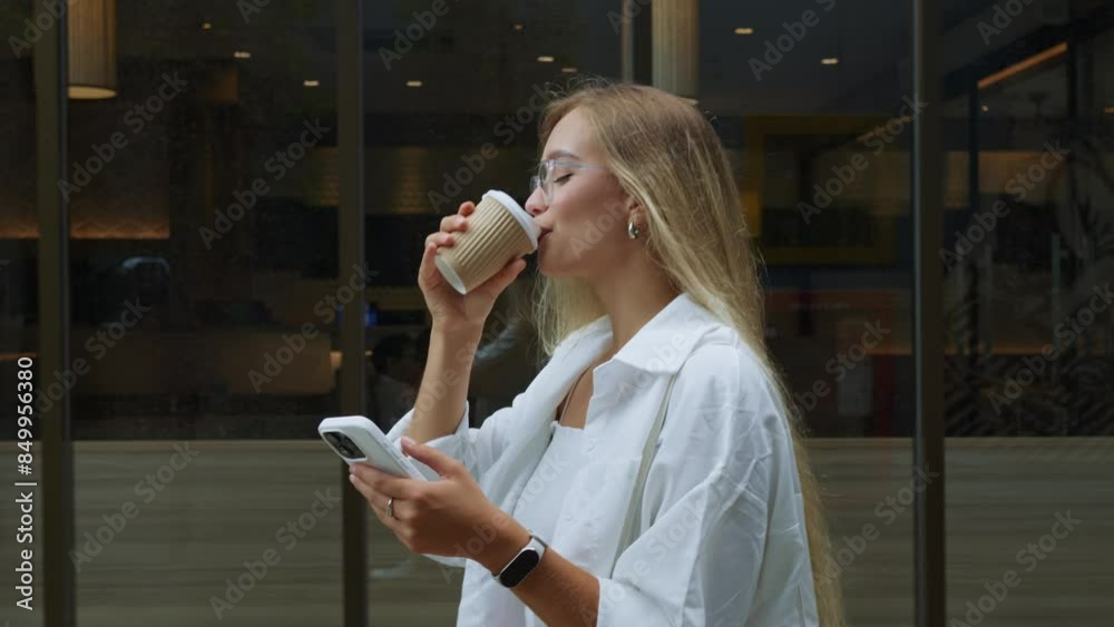 Young stylish blonde smiling woman with glasses walking along street enjoying coffee using mobile phone. Millennial blonde woman in glasses strolls down street sipping coffee texting on phone.
