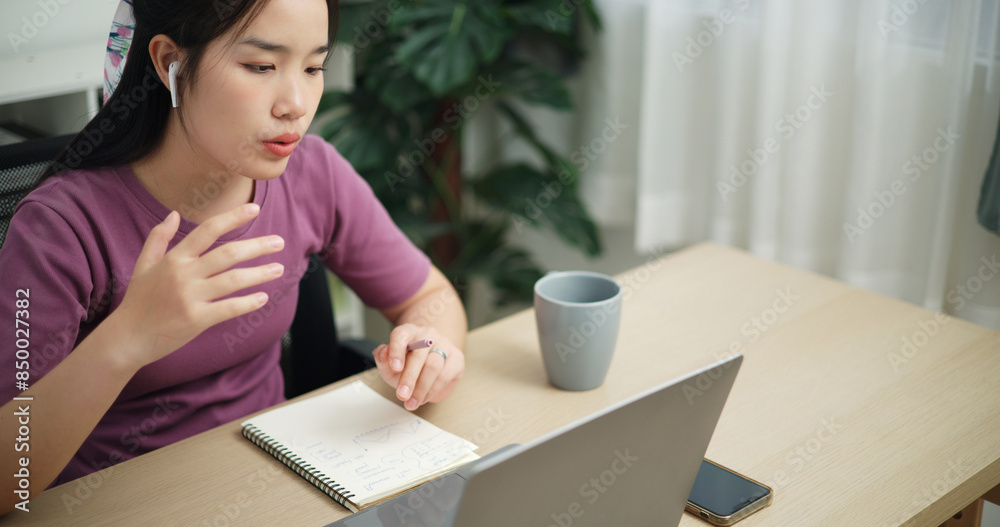 High angle view, Young asian businesswoman wear wireless headphones having a video conference on laptop and writing on note at home office, online meeting for remote work High angle view, Young asian businesswoman wear wireless headphones having a video conference on laptop and writing on note at home office, online meeting for remote work