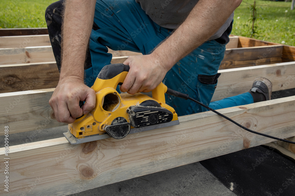 A carpenters hands are shown using a power planer to smooth out a wooden beam. The carpenter is wearing blue pants and is kneeling on the wooden beams. A carpenters hands are shown using a power planer to smooth out a wooden beam. The carpenter is wearing blue pants and is kneeling on the wooden beams.