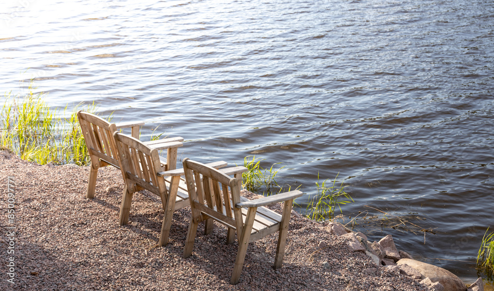 Two wooden chairs sit on a pebbled lakeshore, facing the calm water. The sun casts a golden glow on the scene, creating a peaceful and serene atmosphere. Two wooden chairs sit on a pebbled lakeshore, facing the calm water. The sun casts a golden glow on the scene, creating a peaceful and serene atmosphere.